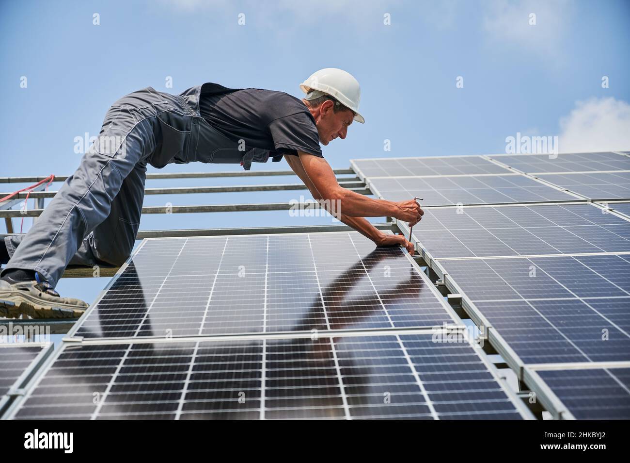 Worker mounting photovoltaic solar panel system outdoors. Man engineer placing solar module on metal rails, wearing construction helmets and work gloves. Renewable and ecological energy. Stock Photo