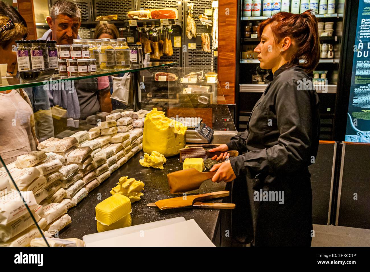 A saleswoman shapes butter with wooden paddles at the counter of Maison du Beurre Bordier in Saint-Malo, Brittany, France, with cured meats and preserves displayed behind glass to her left. An information panel to the right, titled "À l'origine du beurre" ("the origin of butter"), traces the history of butter-making back to antiquity. The shop's façade names 1927 as its founding year; today's Beurre Bordier business goes back to Jean-Yves Bordier, who revived his grandfather's traditional hand-churning method here in 1984. Stock Photo