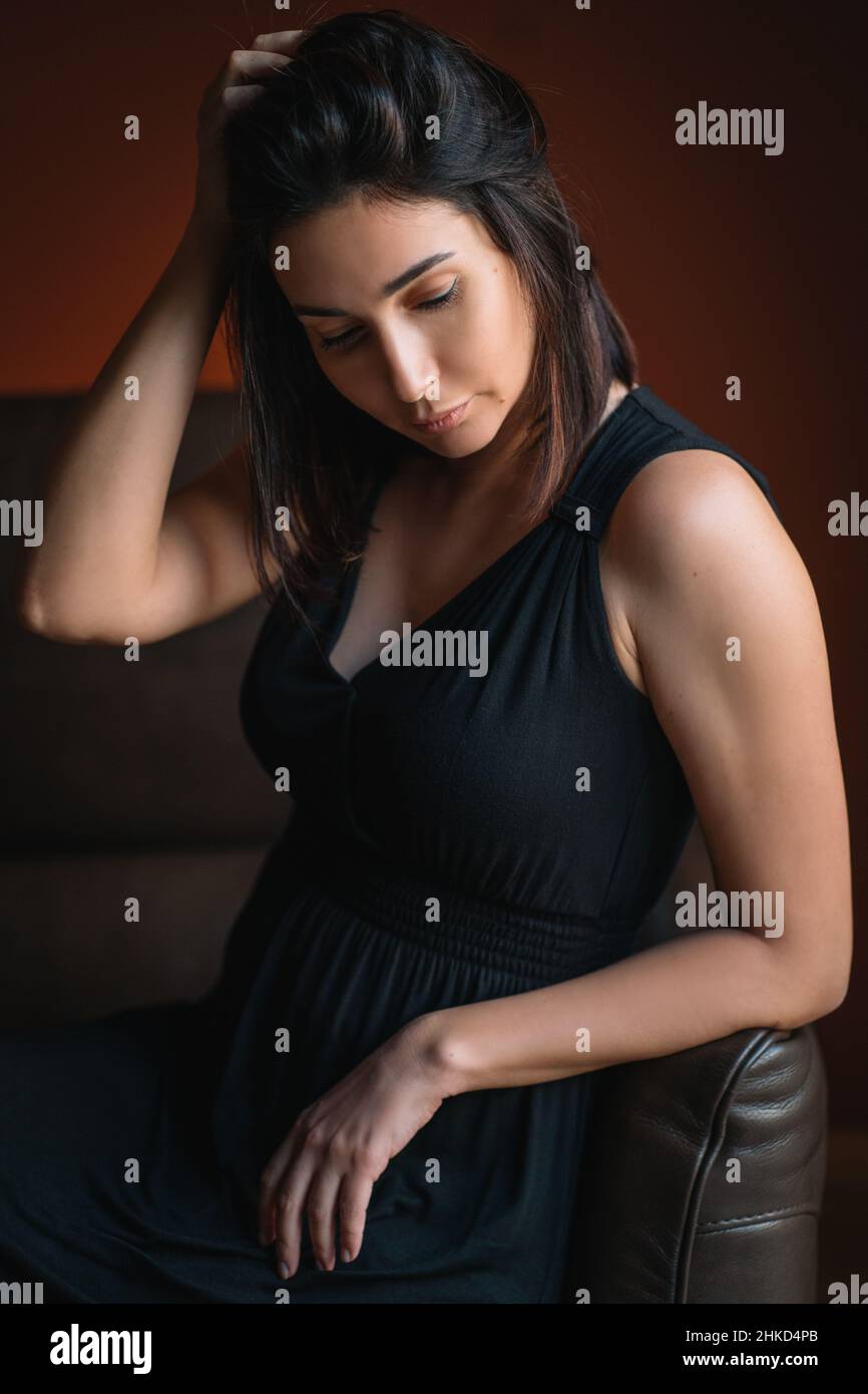 Young beautiful thoughtful woman titivating long dark hair in black dress sit reclining on brown sofa with closed eyes. Stock Photo