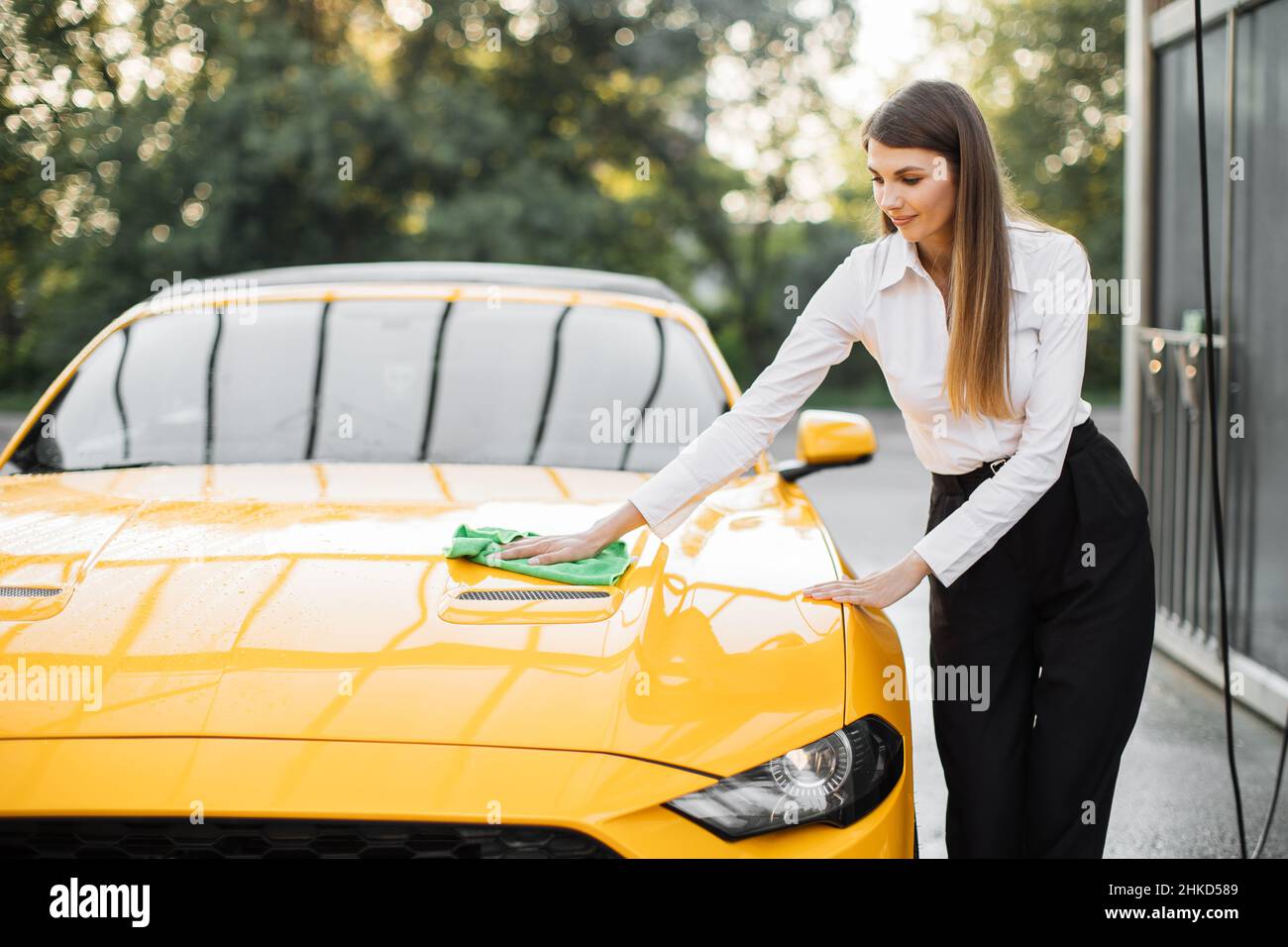 Portrait of a smiling young caucasian businesswoman on white shirt and black pant, cleaning modern sport car hood with green microfiber cloth outdoors, at the self car wash station. Stock Photo
