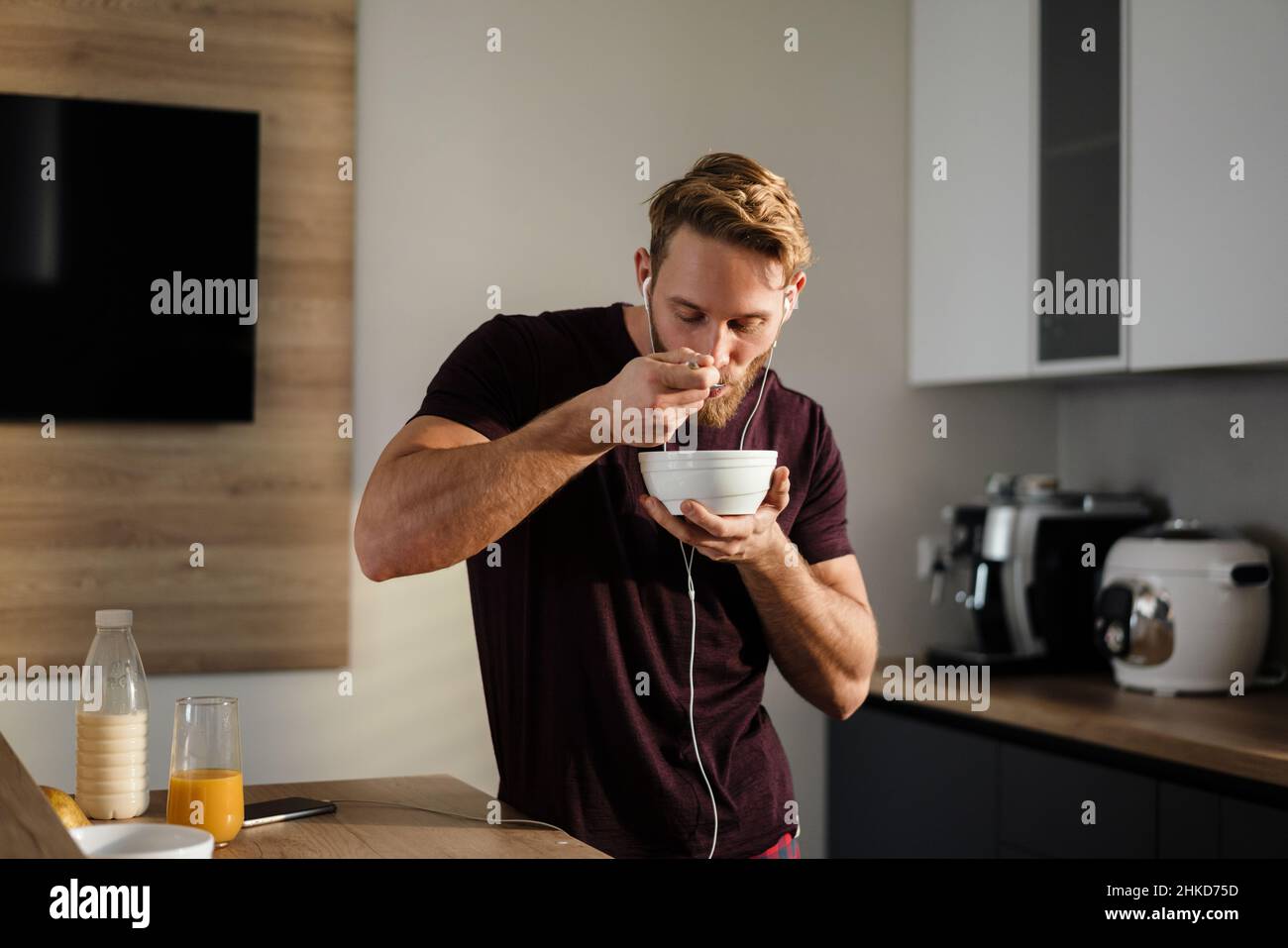 Attractive healthy young man having tasty breakfast while standing in the kitchen, holding bowl, listening to music with earphones Stock Photo
