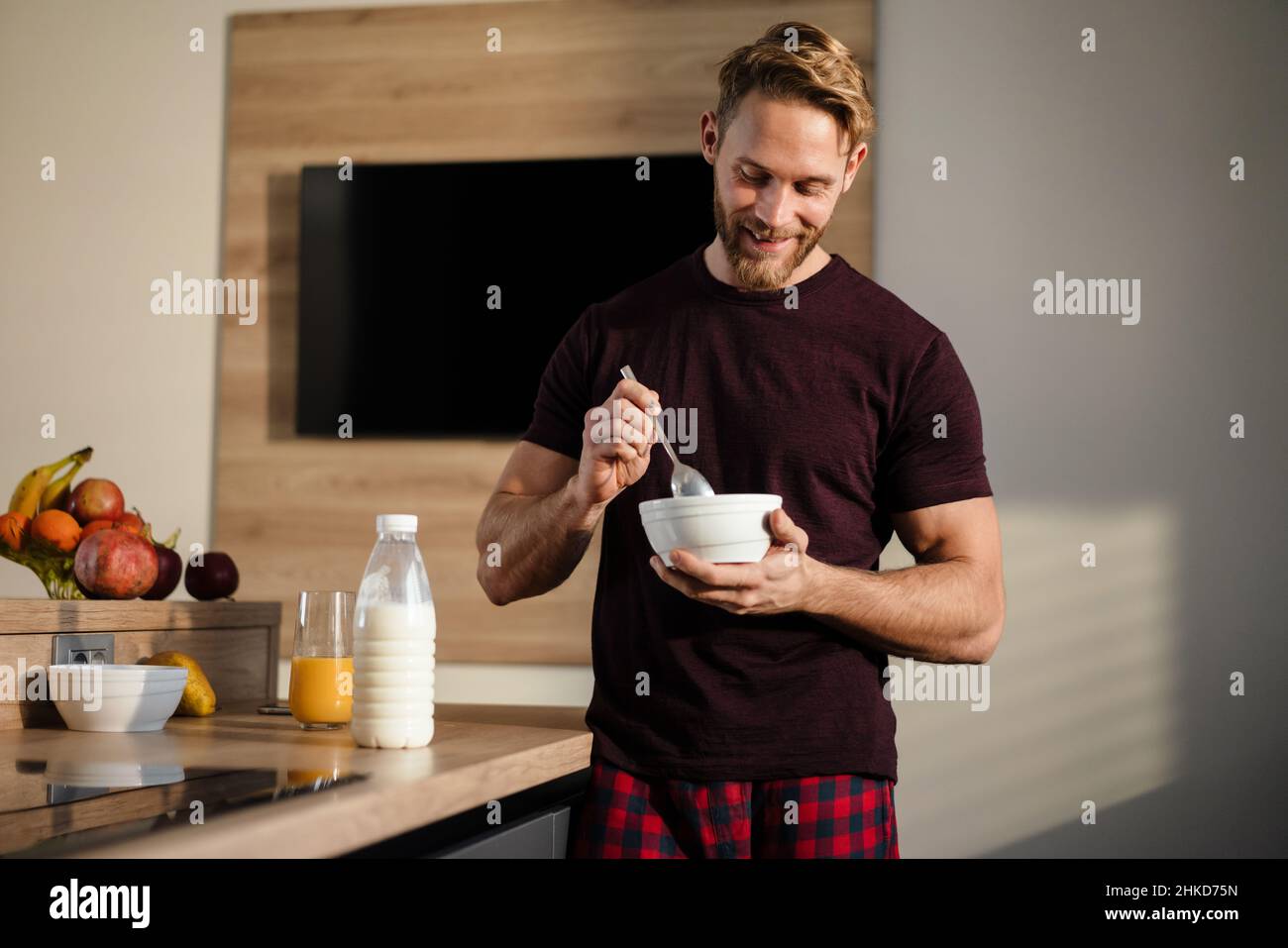 Attractive healthy young man having tasty breakfast while standing in the kitchen, holding bowl Stock Photo