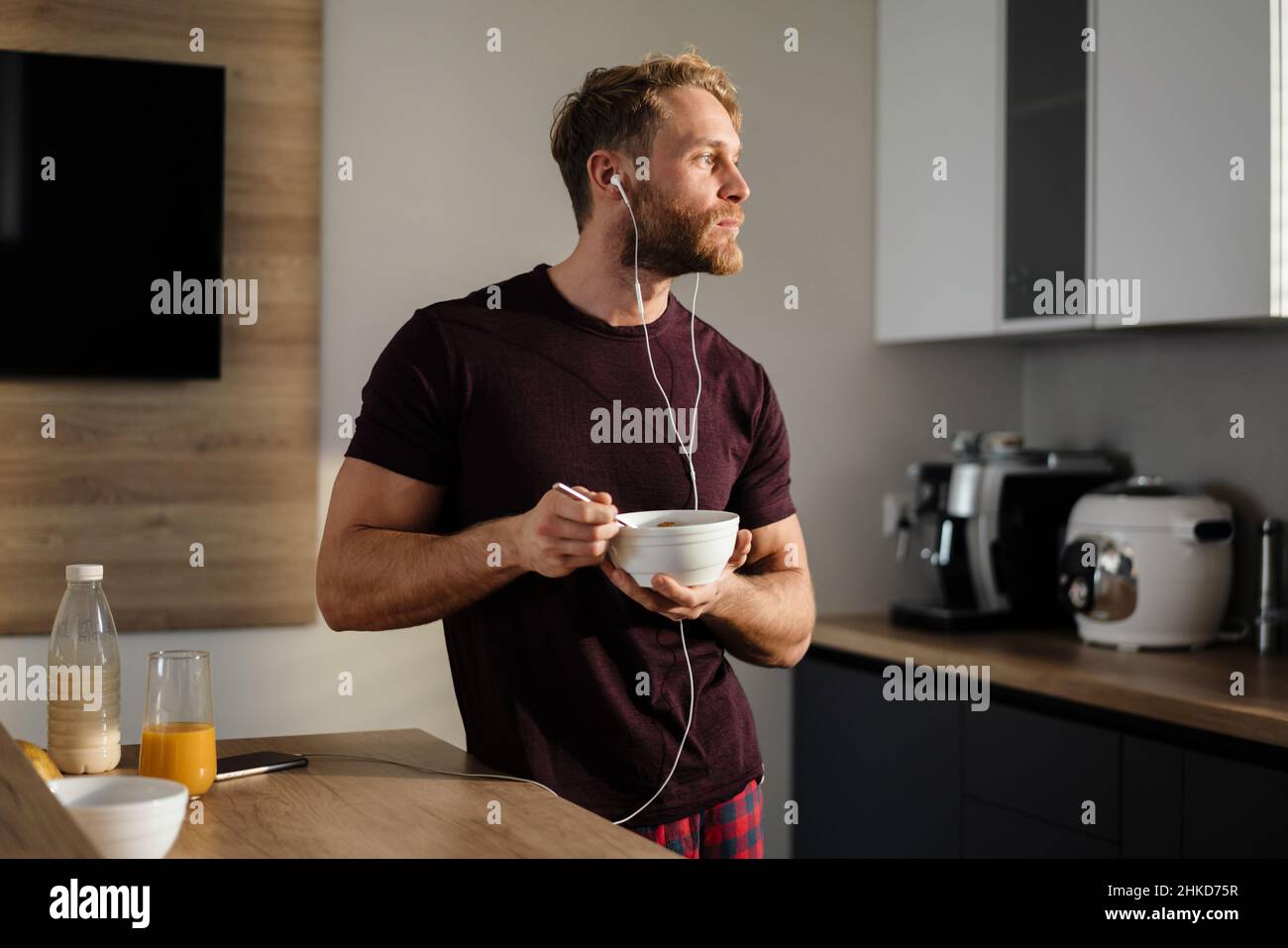 Attractive healthy young man having tasty breakfast while standing in the kitchen, holding bowl, listening to music with earphones Stock Photo