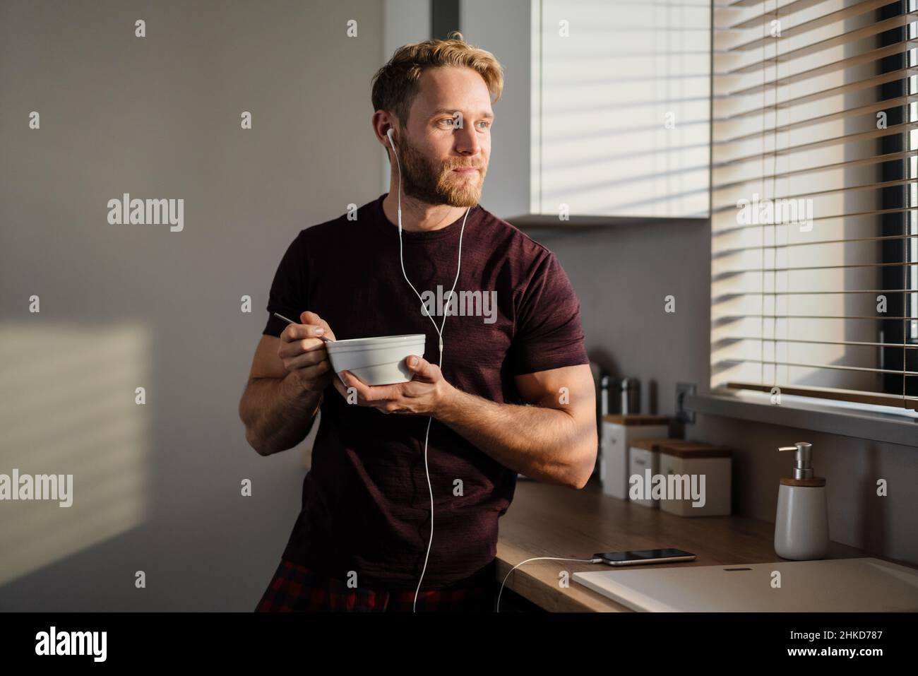 Attractive healthy young man having tasty breakfast while standing in the kitchen, holding bowl, listening to music with earphones Stock Photo