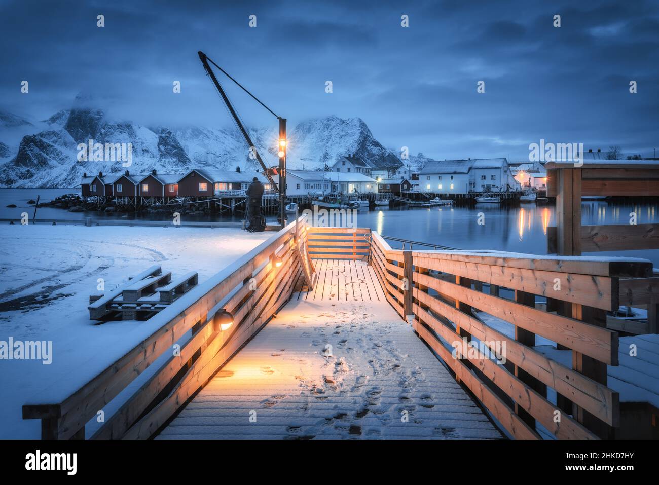 Snowy wooden pier on the sea coast with lights, rorbu, mountains Stock Photo