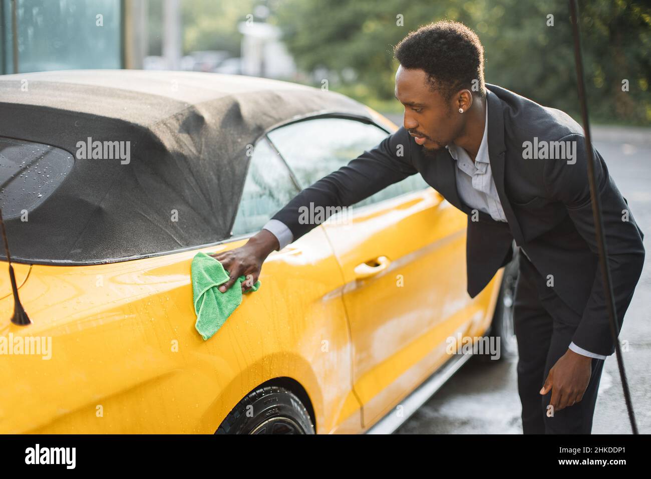 Car washing on open air. Young hipster African bearded man cleaning a  wheel, car rims of modern luxury red car with yellow microfiber cloth, at  outdoor car wash self service - Stock