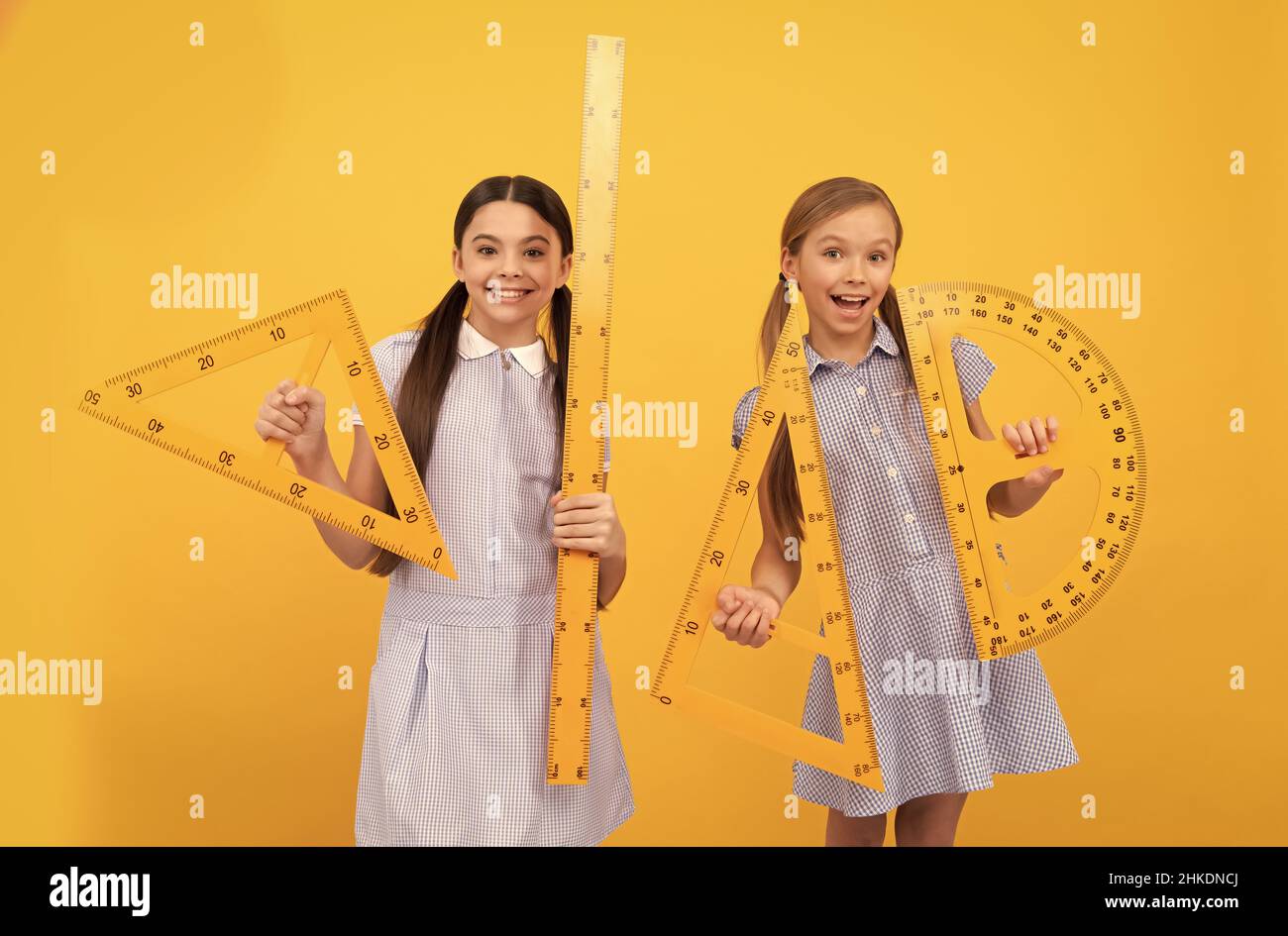Happy school age children hold geometric tools for geometry lesson yellow background, schooling Stock Photo