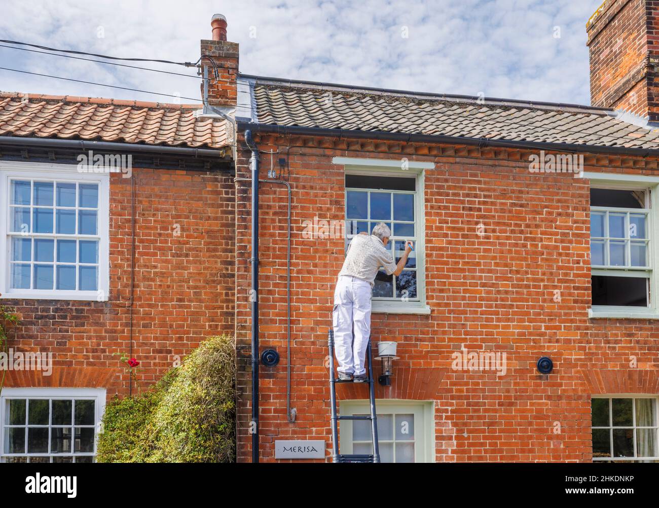 An elderly man on a ladder paints the sash window frame of a house in Cley-Next-The-Sea, a village on the north coast of Norfolk, East Anglia, England Stock Photo