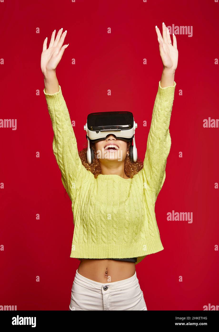 Excited African American female in VR glasses standing with raised arms and exploring cyberspace on red background in studio Stock Photo