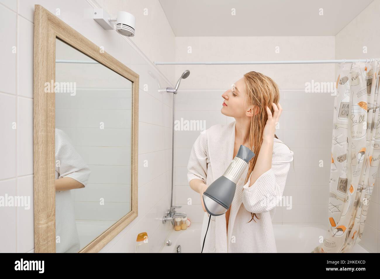 Woman in bathrobe drying her hair with dryer standing in profile in
