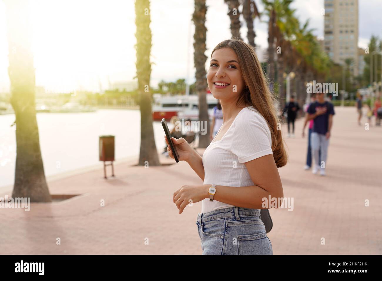 Teenager girl using her cellphone on the waterfront while strolling at sunset Stock Photo