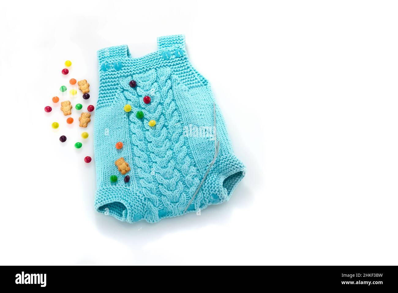 Hand knitted suit for a newborn with an Aran pattern, knitting needles, sweets and bears on a white background. Place for text. Baby blue knitted cash Stock Photo