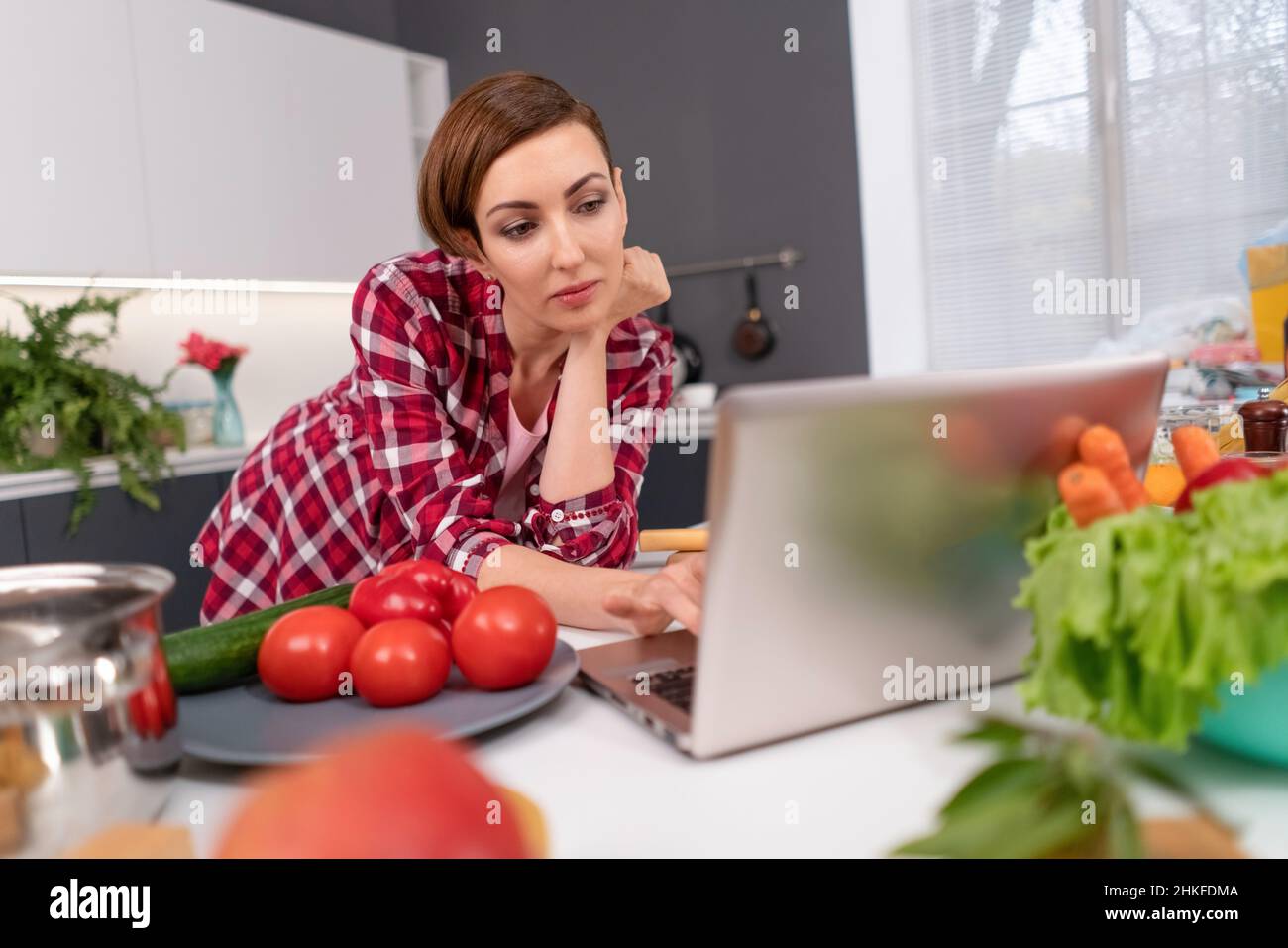 Short haired woman use laptop having video call near fresh vegetables in kitchen. Bored or interested woman uses laptop. Young woman at kitchen searching online for healthy food recipes.  Stock Photo