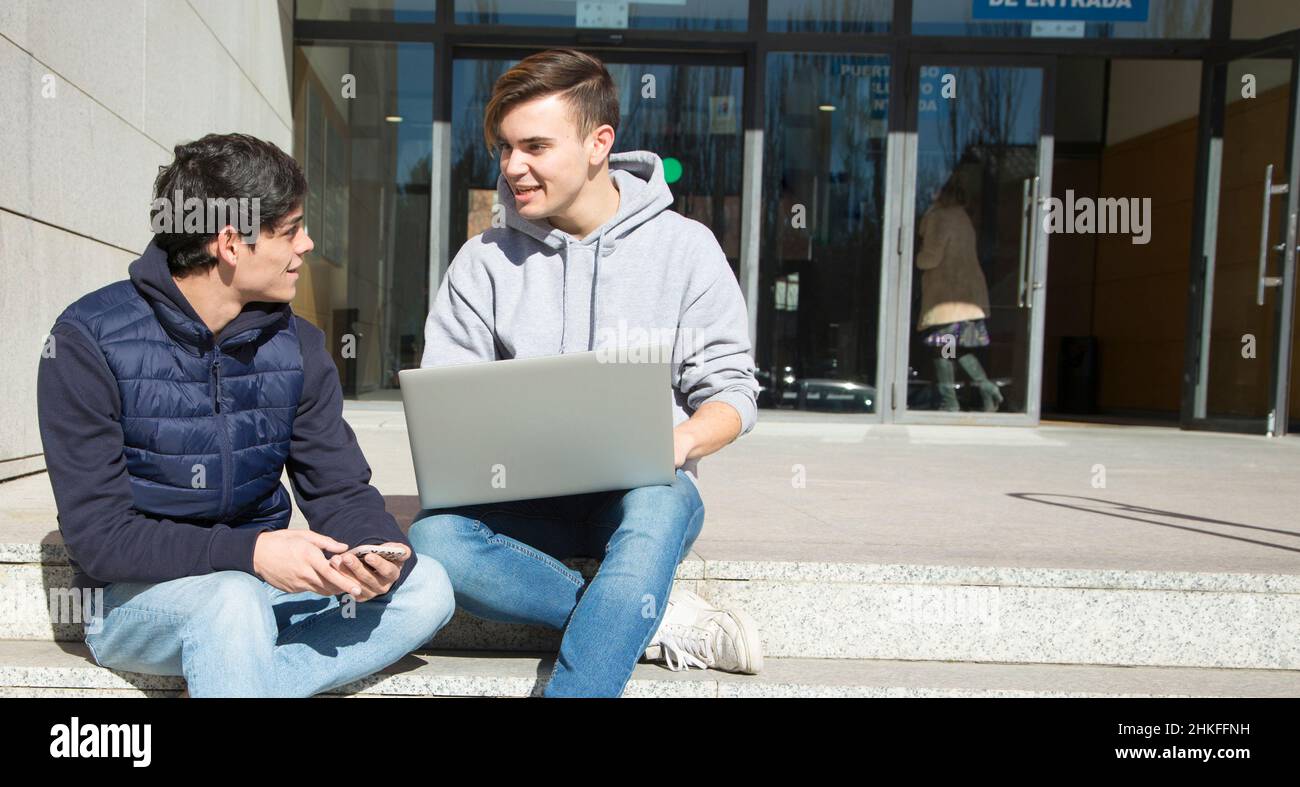Couple two young gay men sitting on stairs with mobile phone and laptop looking at each other and smiling. Stock Photo