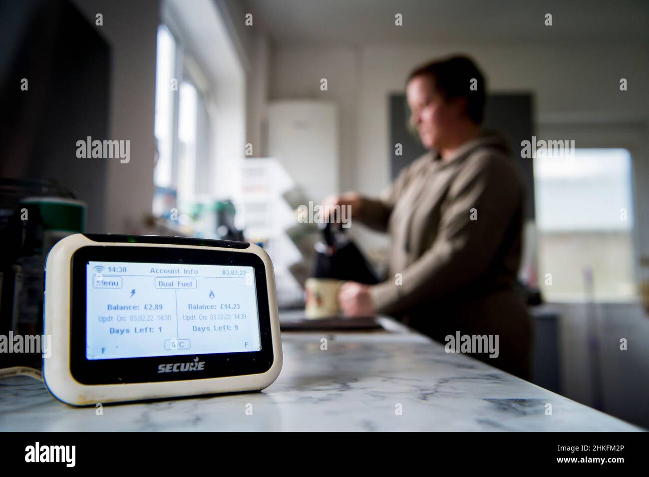A UK prepaid energy meter in the foreground with a woman in the background boiling a kettle and having a cup of tea.The energy crisis is leading to hu Stock Photo