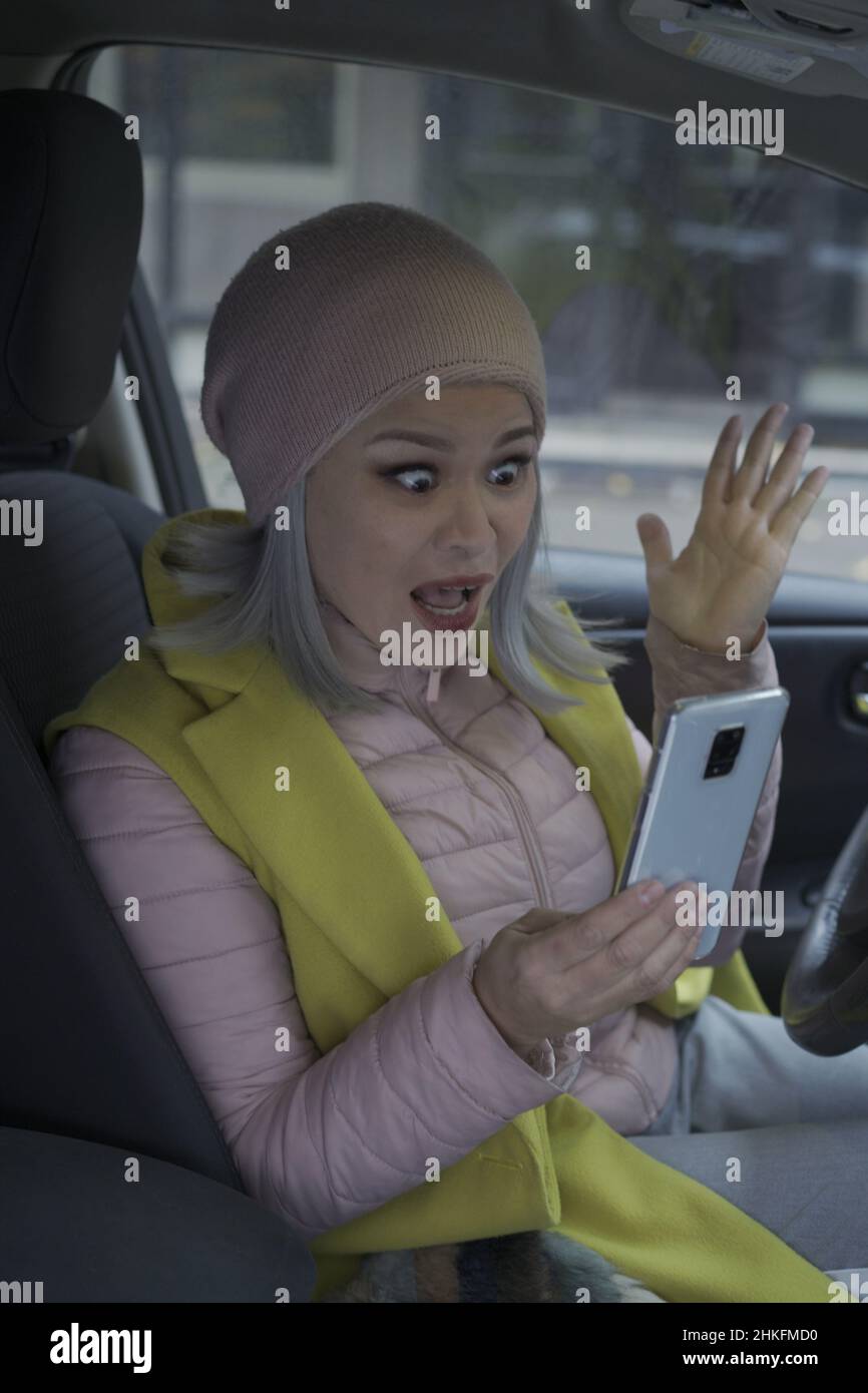 Shocked middle aged asian blond woman in the car. Asian woman use mobile phone sitting and driver seat in the car. Gray haired middle aged asian woman in the car.  Stock Photo