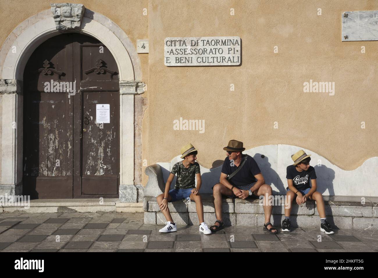 Italy, Sicily, Taormina, father and his two sons wearing straw hats on the stone bench of the Sant'agostino church on the 9-Avril square Stock Photo