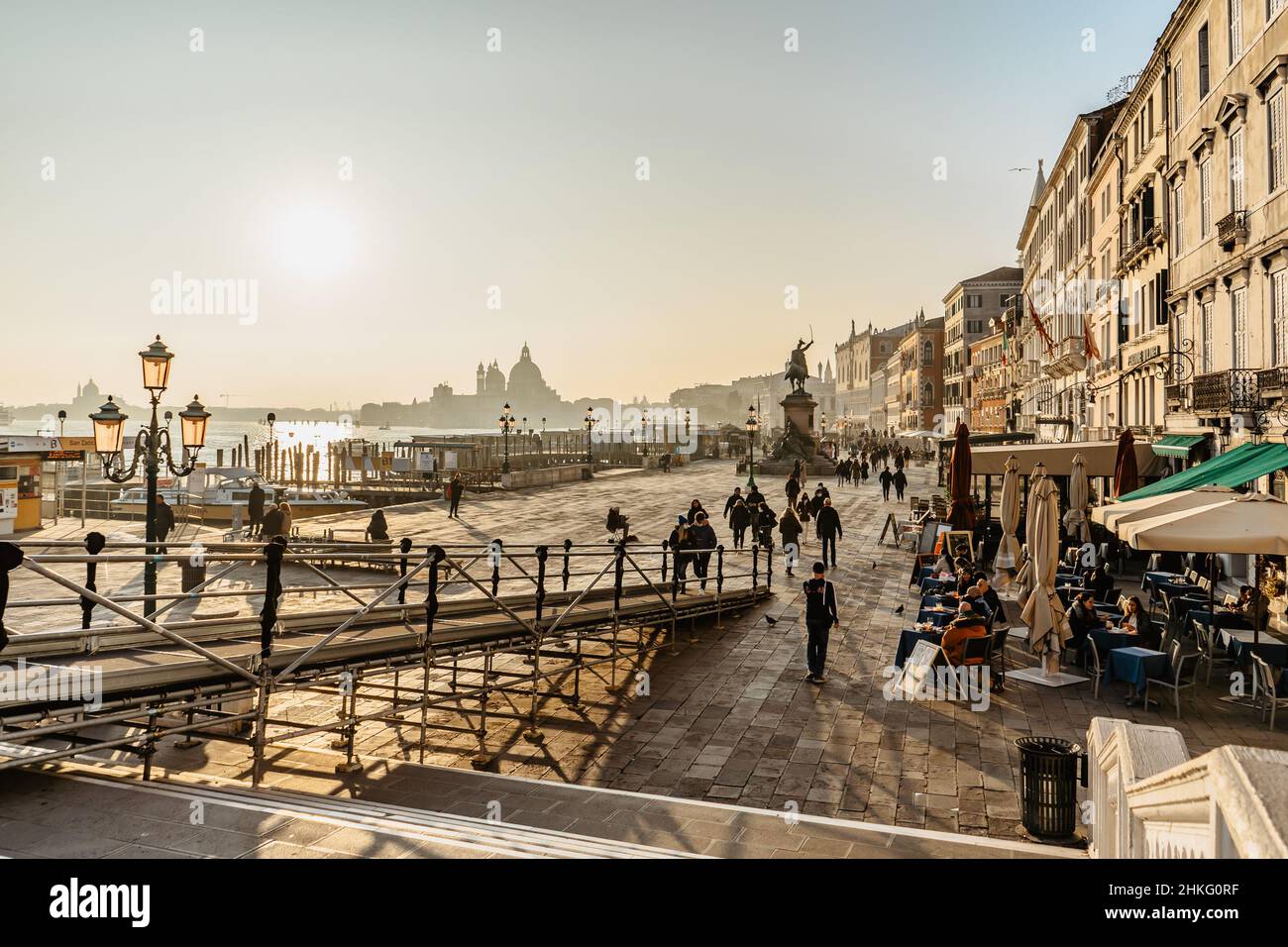 Venice,Italy-January 28,2022.People walking on embankment,tourist sitting in restaurant on sunny day,church,canal and gondolas in background.Venetian Stock Photo