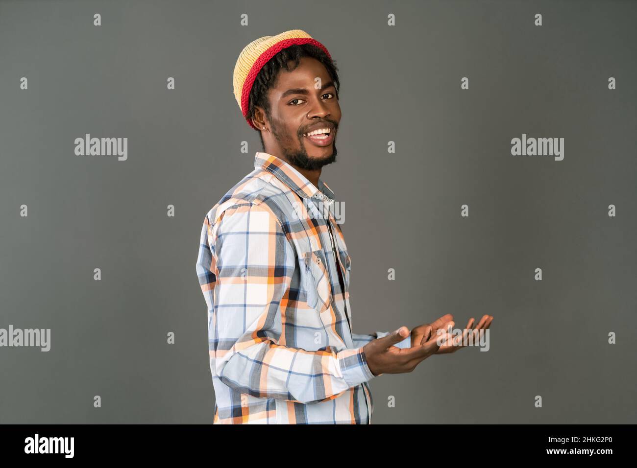 Young black man in afro hat smiling happy gesturing with open hands half lifted up. Cheerful african american young man in plaid shirt looking at camera isolated on gray background.  Stock Photo