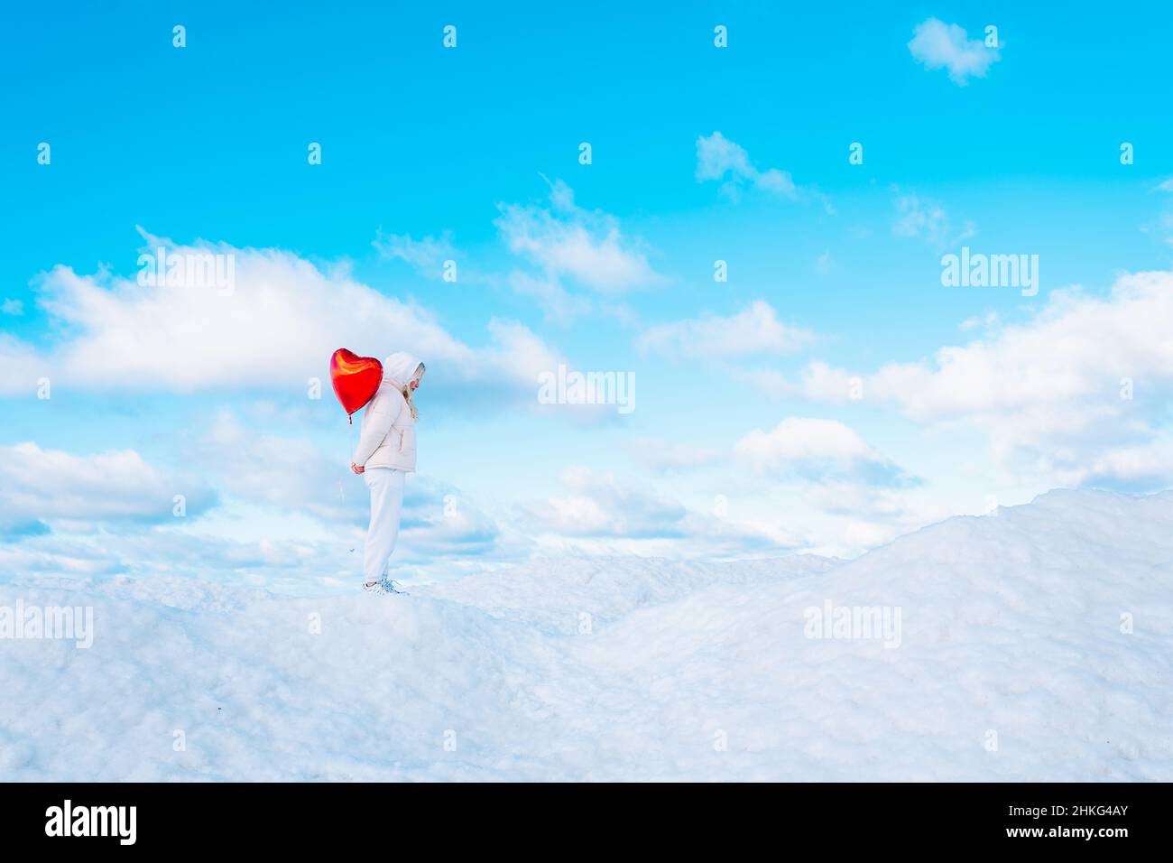 Portrait of young woman in white clothes with red heart balloon walking on snow cover with blue cloudy sky on background Stock Photo