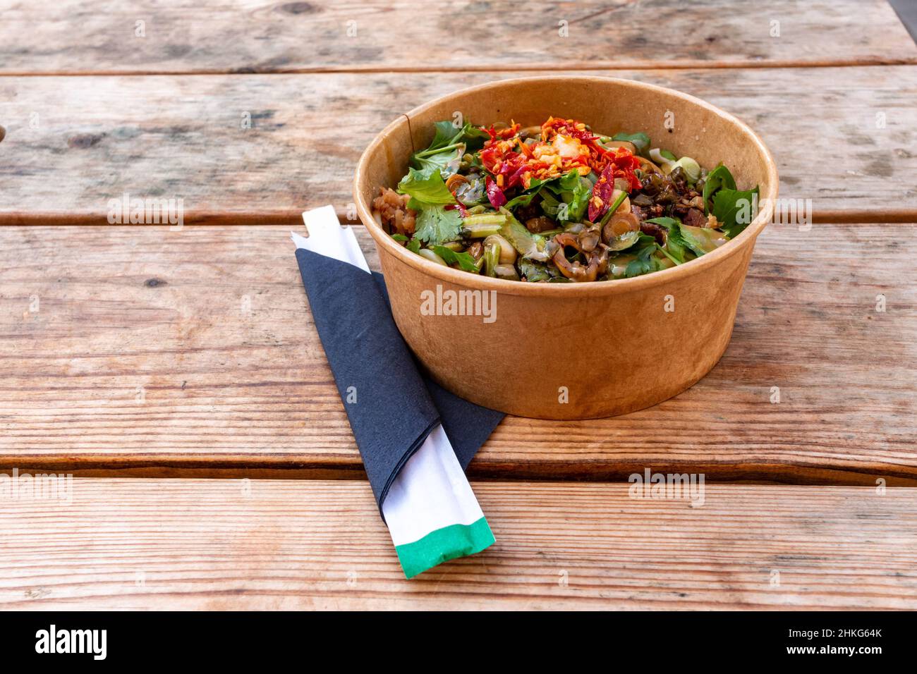 A bowl of hot dry noodles with chilies and coriander topping sits next to disposable chopsticks. Streetfood available from the Market in Cambridge, UK. Stock Photo