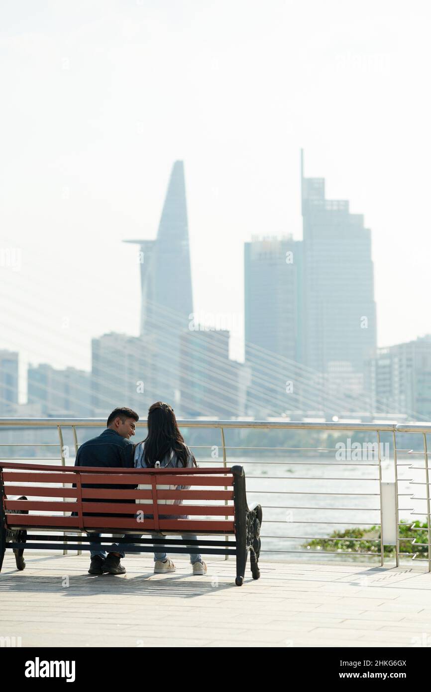 Happy young couple sitting on bench on bridge, talking and enjoying city view Stock Photo