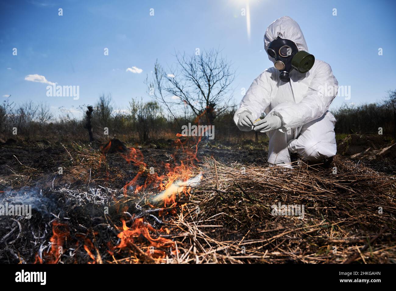 Unrecognizable person burning old dried grass in field. Focus on a fire. Ecologist holding matchbox and setting fire to dry grass under blue sky, wearing protective radiation suit and respirator. Stock Photo