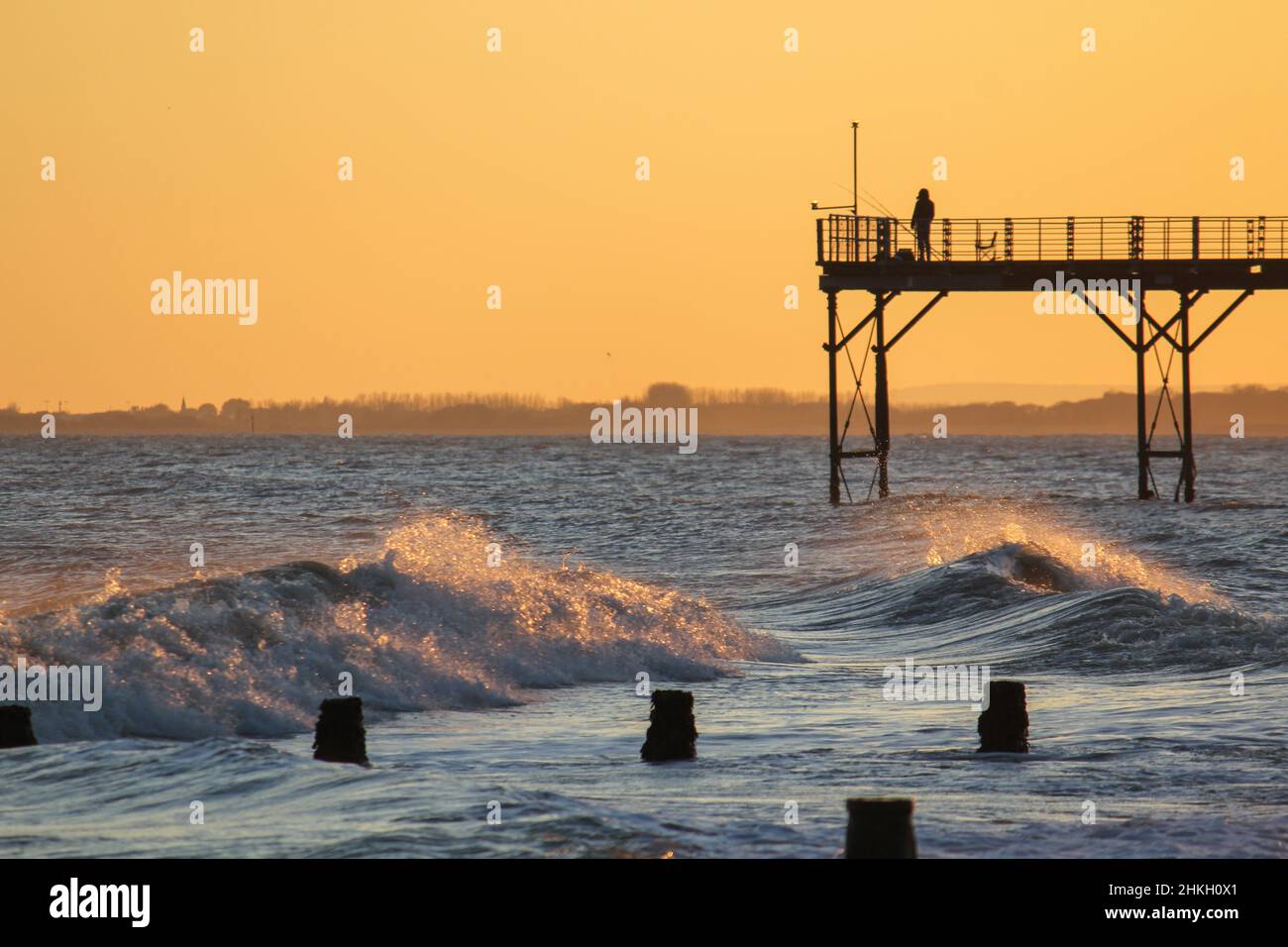 A lone fisherman with fishing rod standing on the end of Bognor Regis pier at sunset with waves breaking on the shore. Stock Photo
