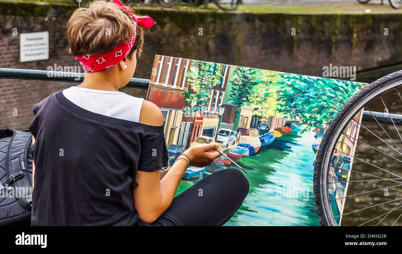 Eine junge Frau im Schneidersitz und rotem Stirnband auf einer Brücke in Amsterdam malt eine typische Häuserfront an einer Gracht Stock Photo