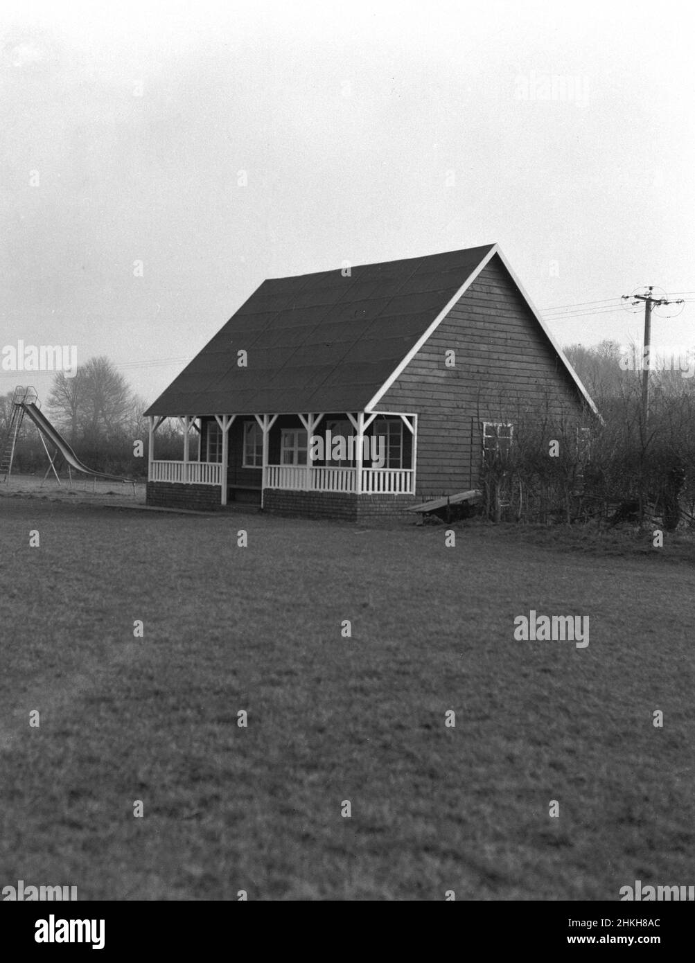 1950s, historical, exterior view of a traditional sports pavilion of the era built with a timber frame on a brick base and a high sloping roof. Architecturally distinctive with a covered porch area or verandah at the front, these types of single-storey buildings on the edge of sports fields were seen at many recreational and sports grounds in Britain at this time, providing changing rooms for team sports such as cricket, rugby and football. Beside this newly constructed pavilion at Witney, Oxford, England, UK, a chlldrens play area, with medal slide. Stock Photo