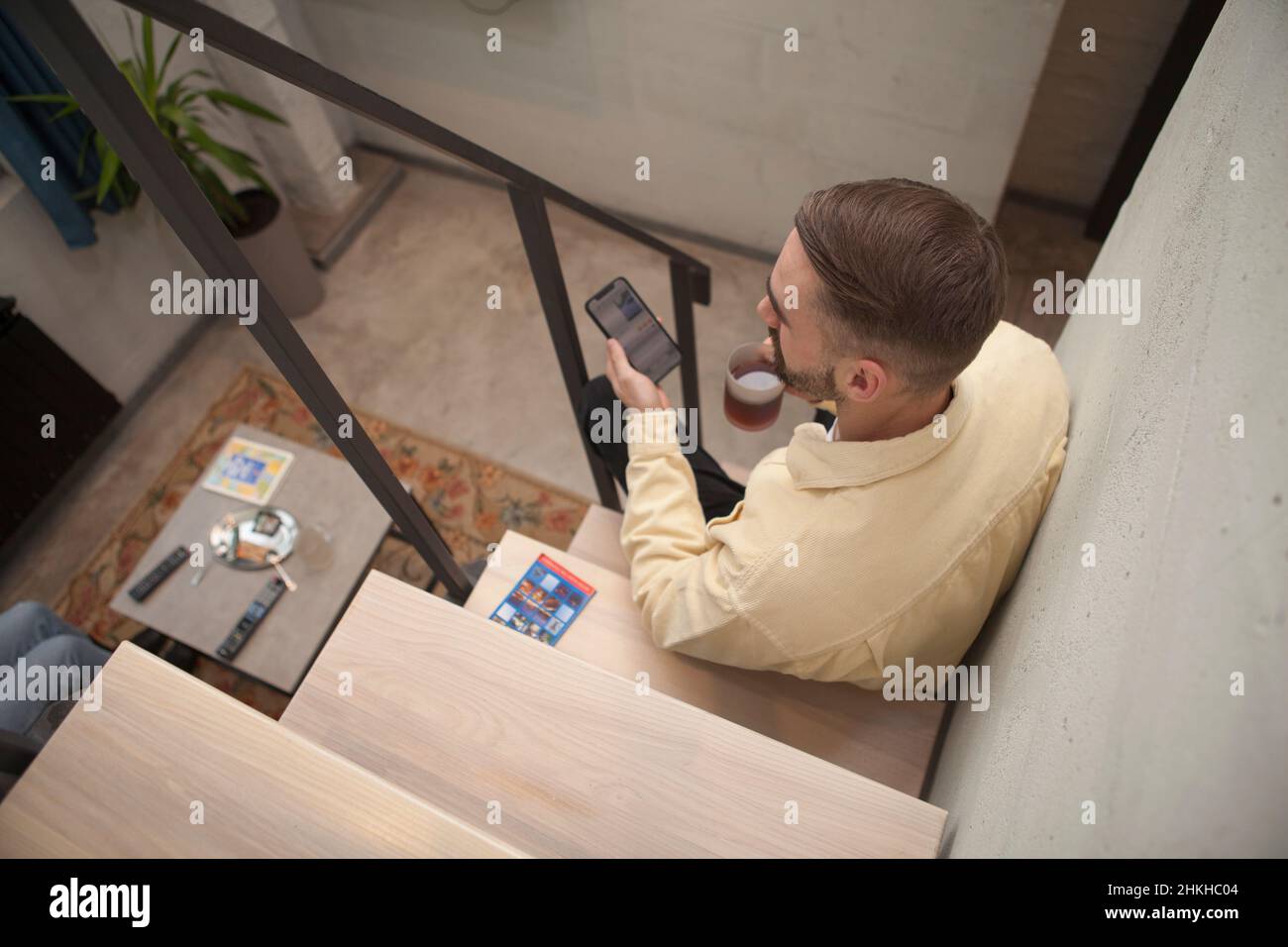 Top view shot of a man having tea while texting on his smart phone, sitting on staircase indoors Stock Photo