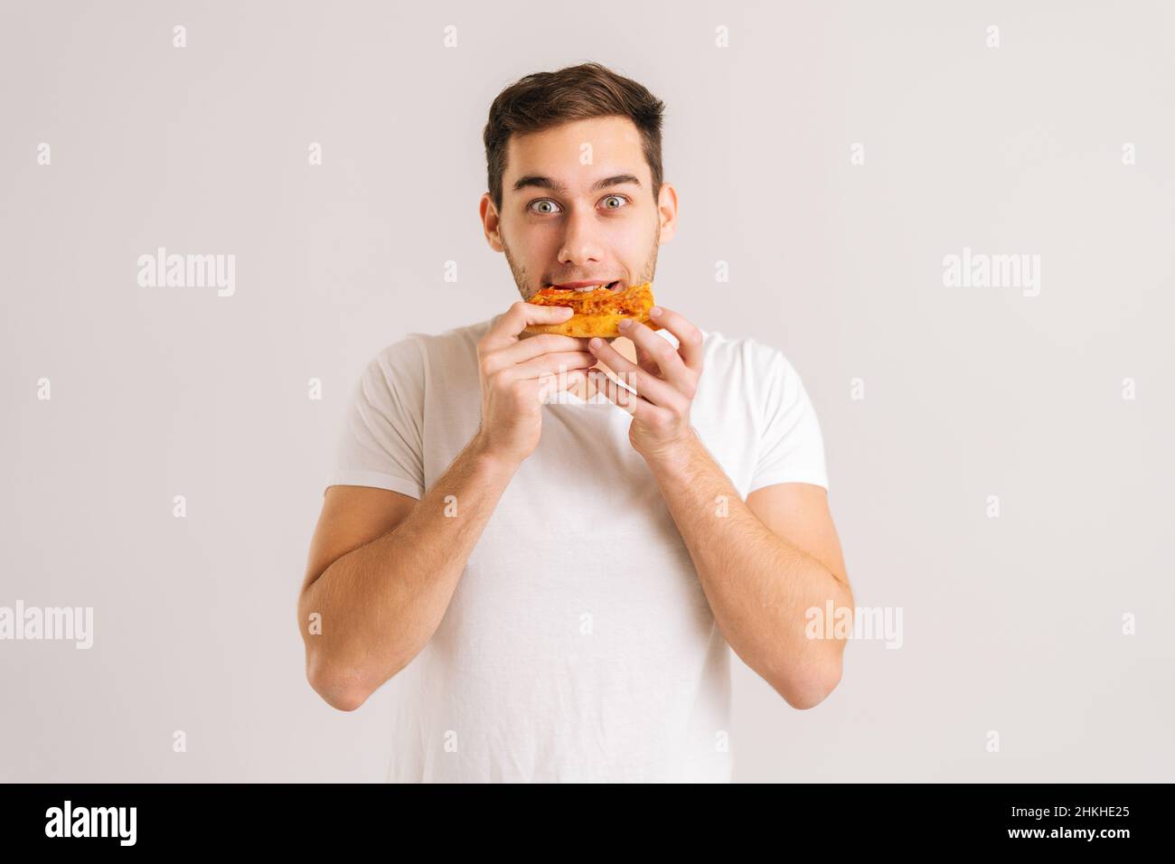 Portrait of happy young man bitting delicious slice of pizza looking at camera on white isolated background. Stock Photo