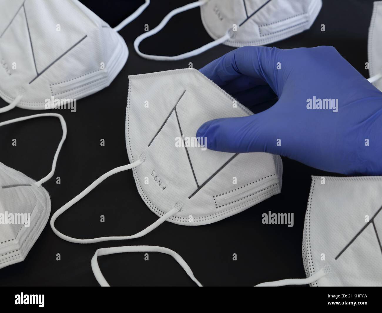 A hand wearing blue nitrile rubber gloves takes a mask from a stockpile of KN95 protective dust masks. Stock Photo