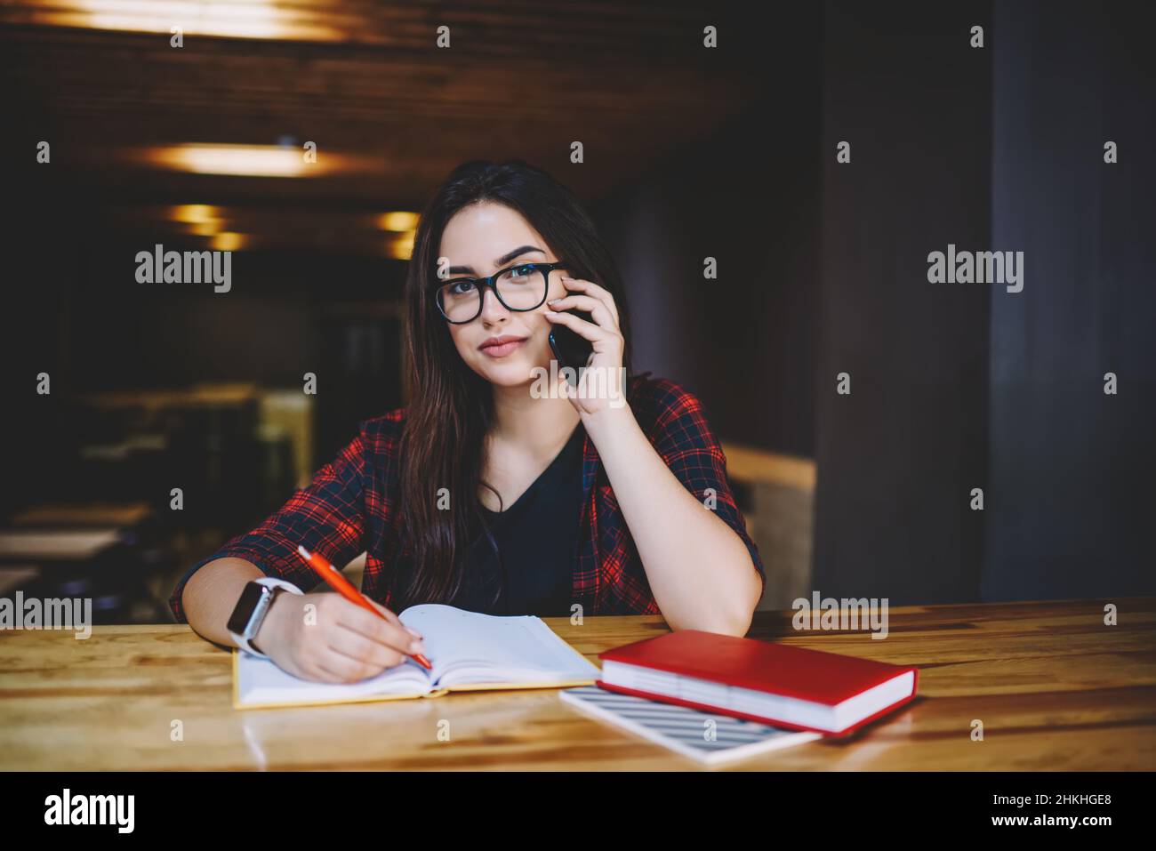 Calm female freelancer calling by smartphone and taking notes in cafe Stock Photo