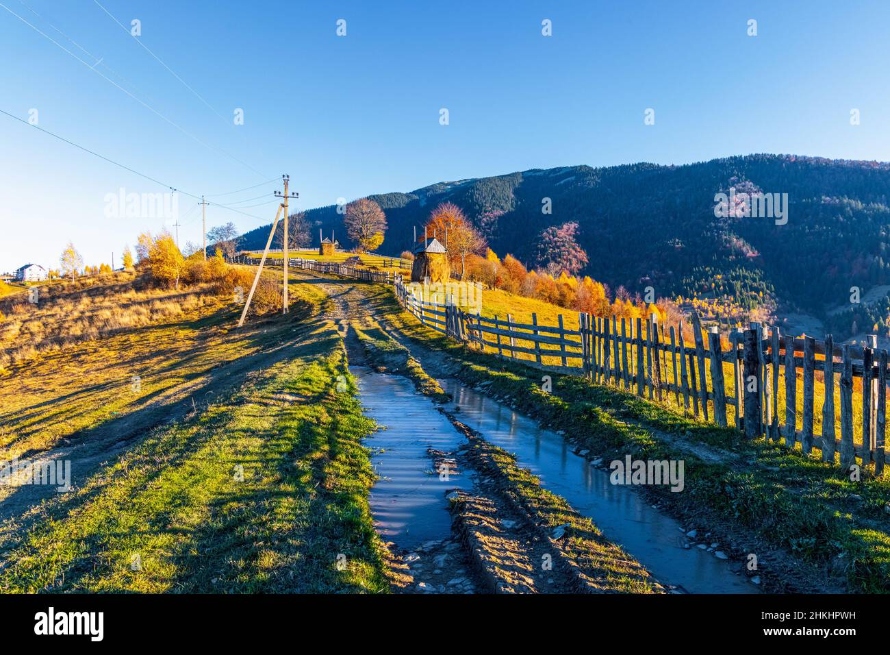 Ground road with dirty puddles after rain runs along wooden fence near highland village against forestry mountains on sunny autumn day Stock Photo