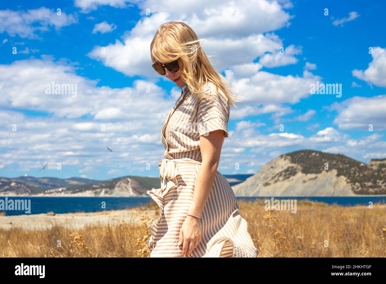 A young blonde woman walks in nature among the sea and mountains on a summer sunny day. Travel and tourism. Stock Photo