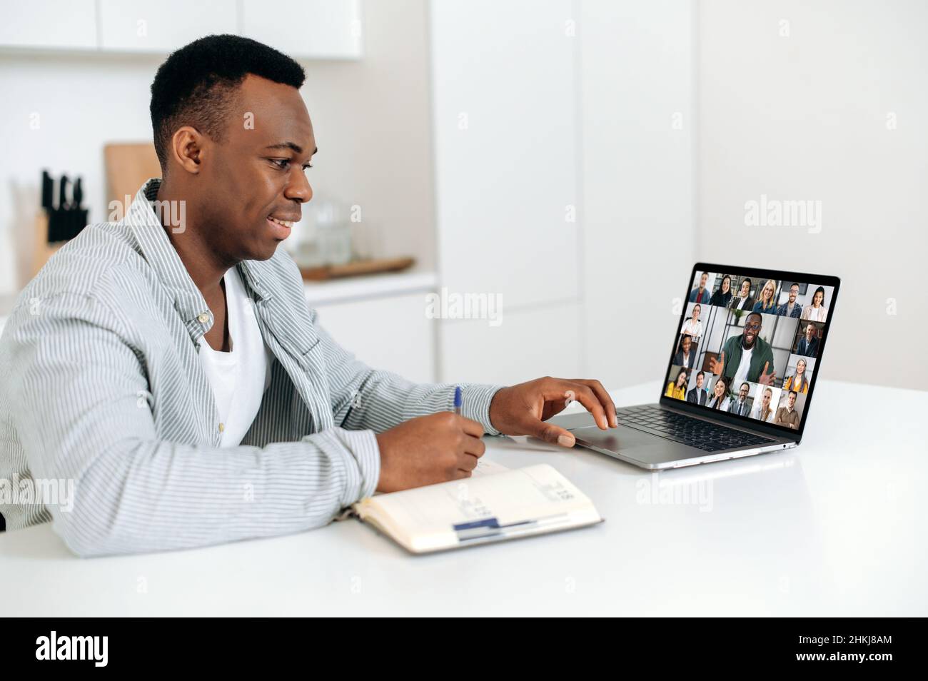 Smart positive african american man sits at a table at home, using a laptop, takes part in a multi-rice video conference with colleagues or employees, takes notes. E-learning concept. Work from home Stock Photo