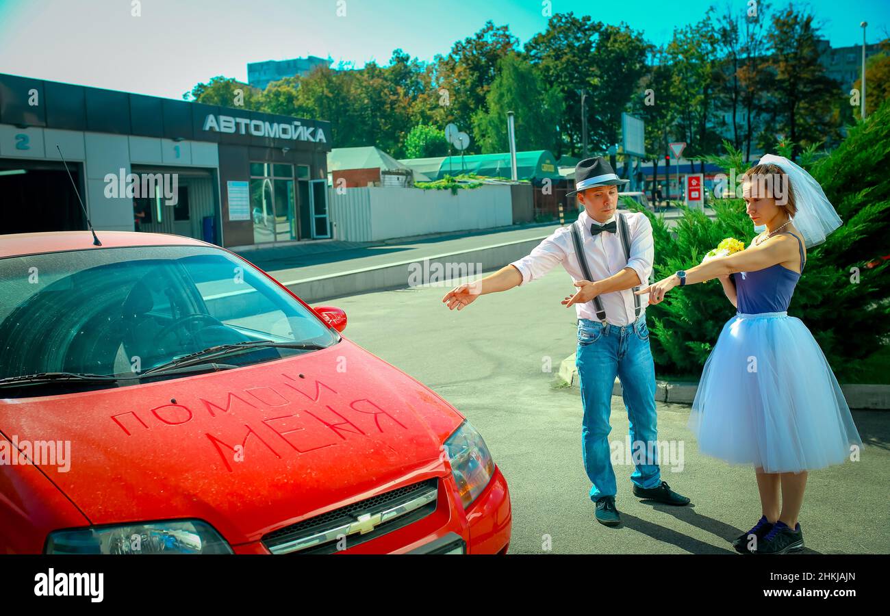 A young man in a hat and a woman with a bouquet of flowers near the car. On the hood in Russian is written 'wash me.' The girl shows that the car need Stock Photo