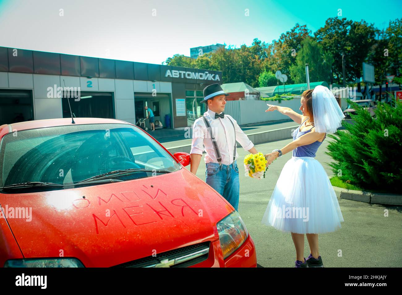 A young man in a hat and a woman with a bouquet of flowers near the car. On the hood in Russian is written 'wash me.' The girl shows that the car need Stock Photo