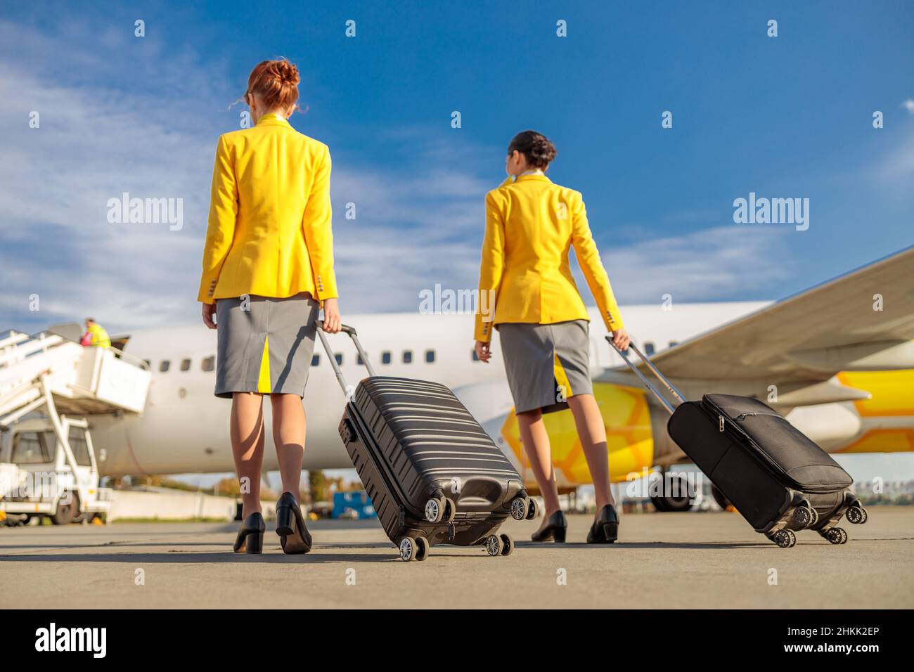Back view of two women flight attendants carrying trolley luggage bags while walking down airfield Stock Photo