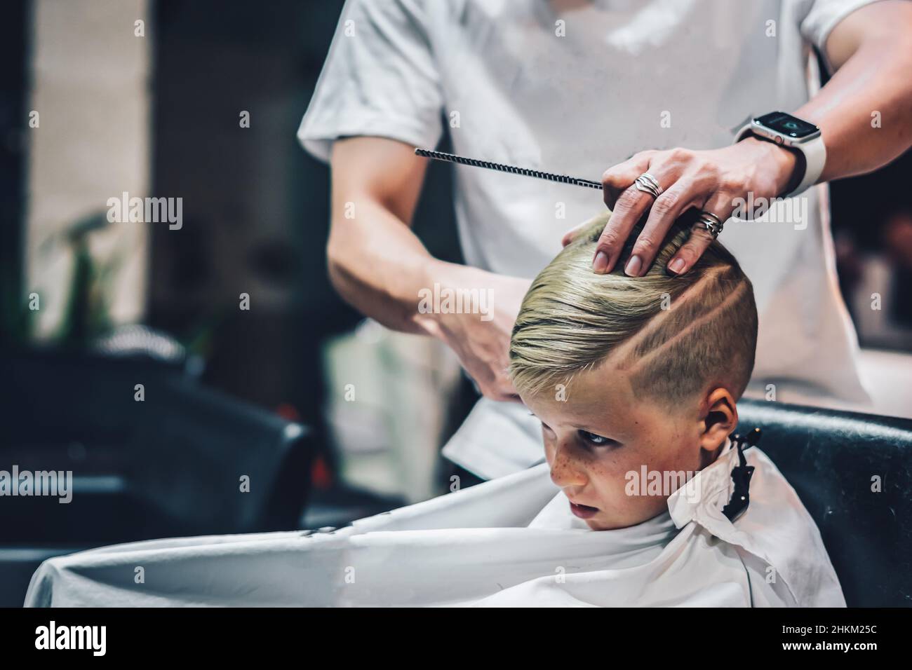 Lifestyle photo Barber shop serious blond boy customer side view.  Hairdresser man work move hand comb tool white cape. Modern style kid short hair  cut Stock Photo - Alamy