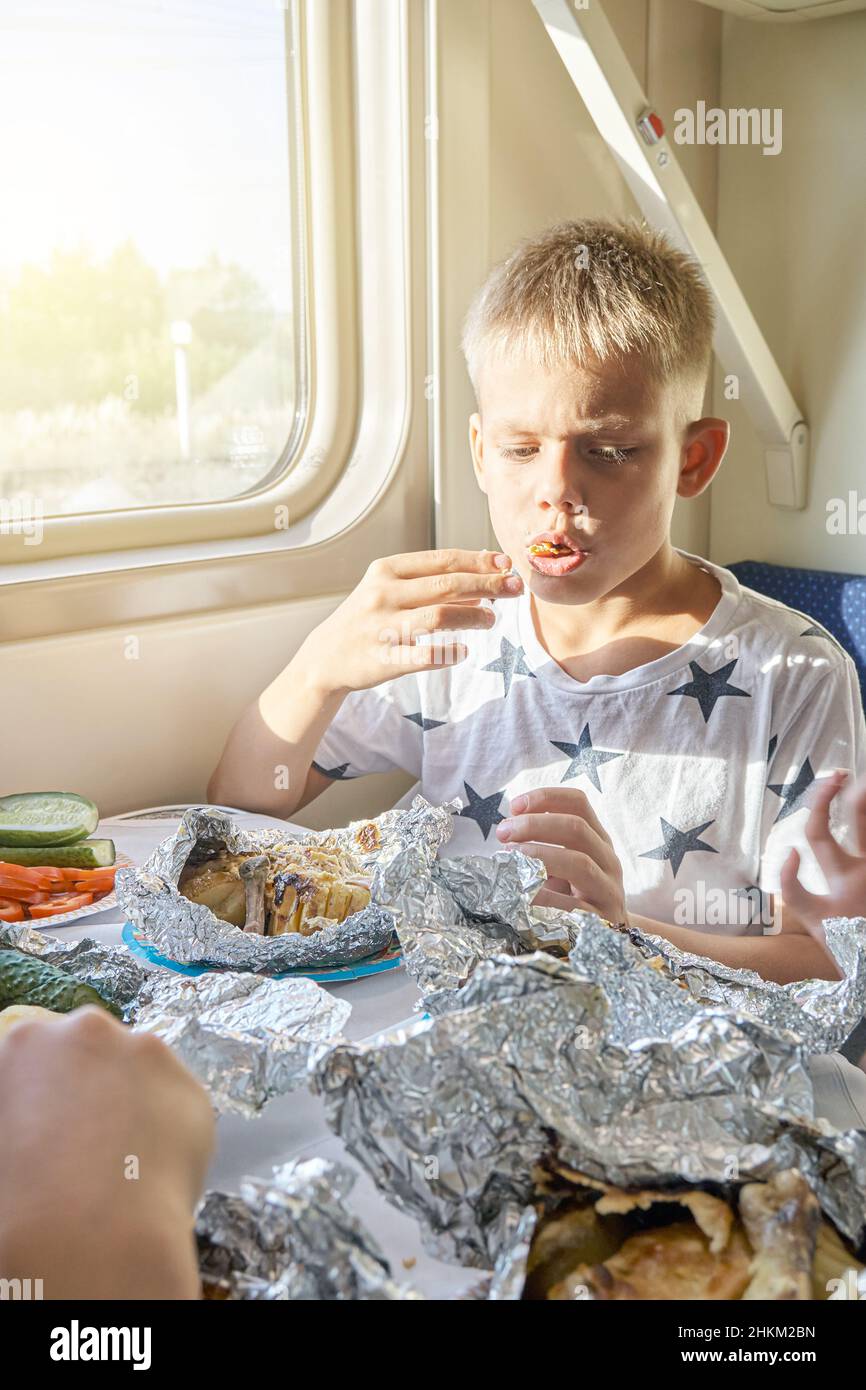 Blond teen boy have lunch eating fried chicken sitting at table in train car against landscape outside window closeup Stock Photo