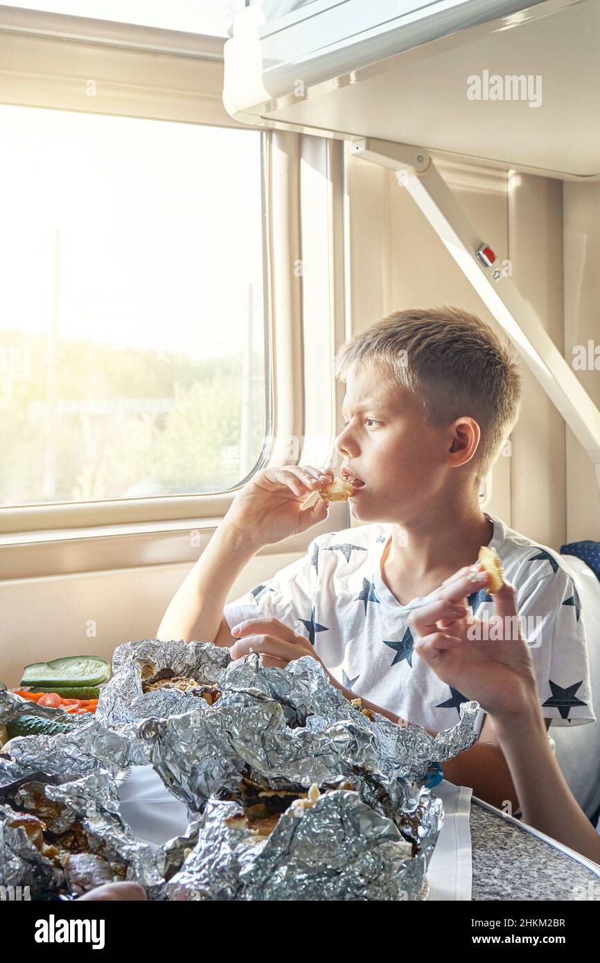 Blond teen boy have lunch eating fried chicken sitting at table in train car against landscape outside window closeup Stock Photo