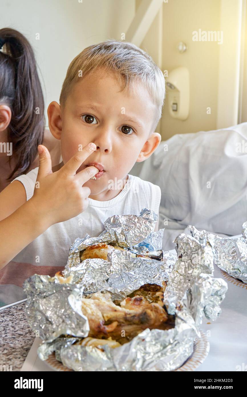 Blond little boy in mother arms licks fingers eating lunch with mouth full of fried chicken lying on foil on table of train wagon closeup Stock Photo