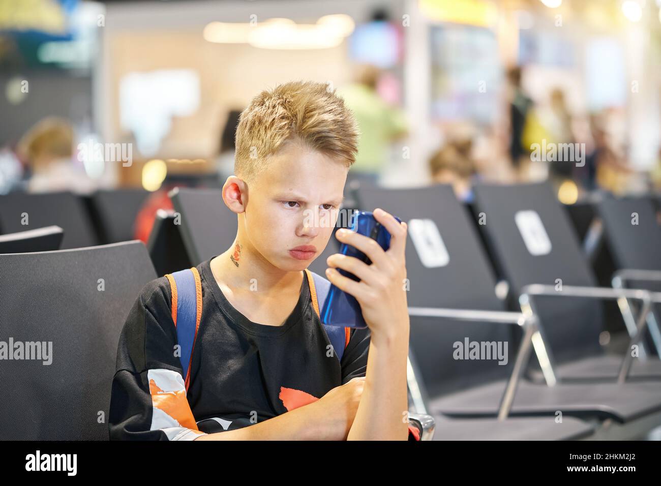 Teenager boy with short blond hairstyle and backpack frowns with serious face expression and reads ebook via phone in airport Stock Photo