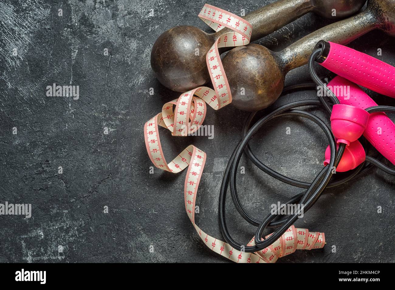 Bright sports concept. Sports equipment as dumbbells, expander with pink handles and centimeter tape on a dark black cement background, top view with Stock Photo