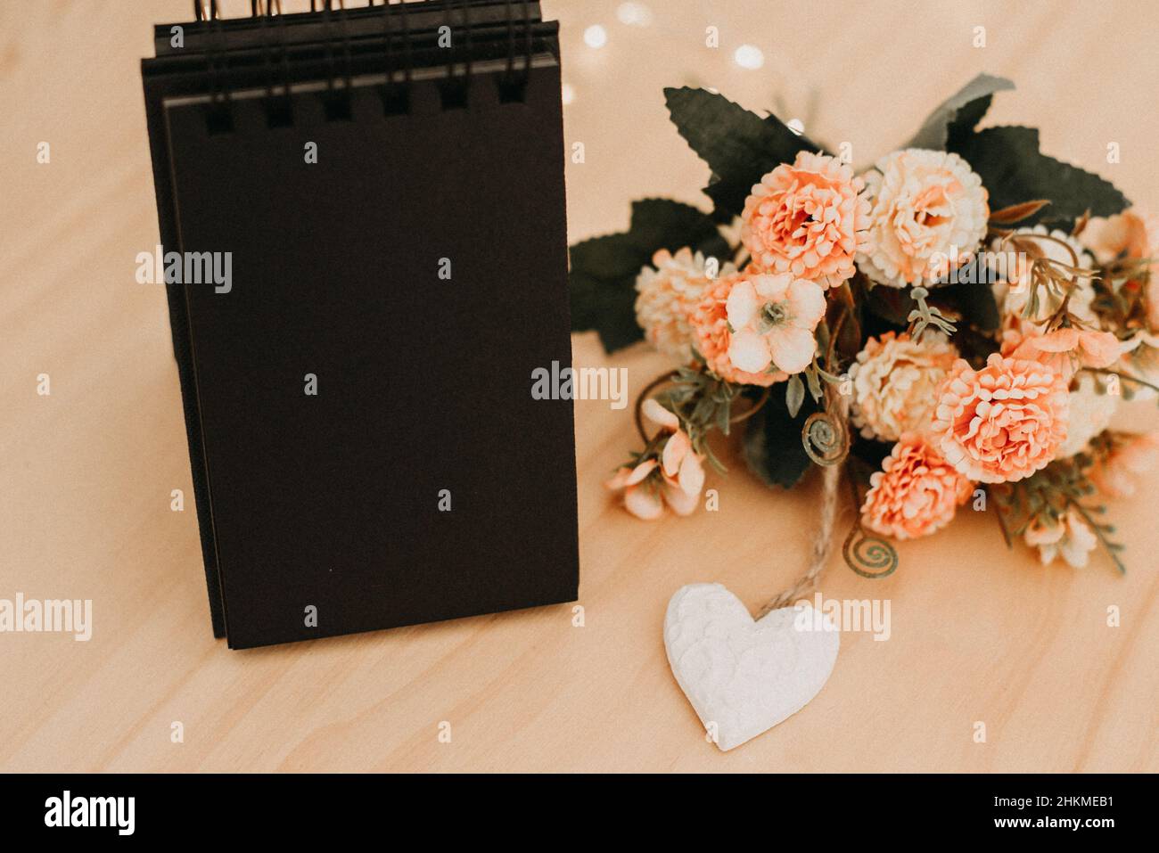 notebook with black sheets with copy space, on a wooden table with a bouquet of flowers and a white heart and bokeh lights around it. Stock Photo
