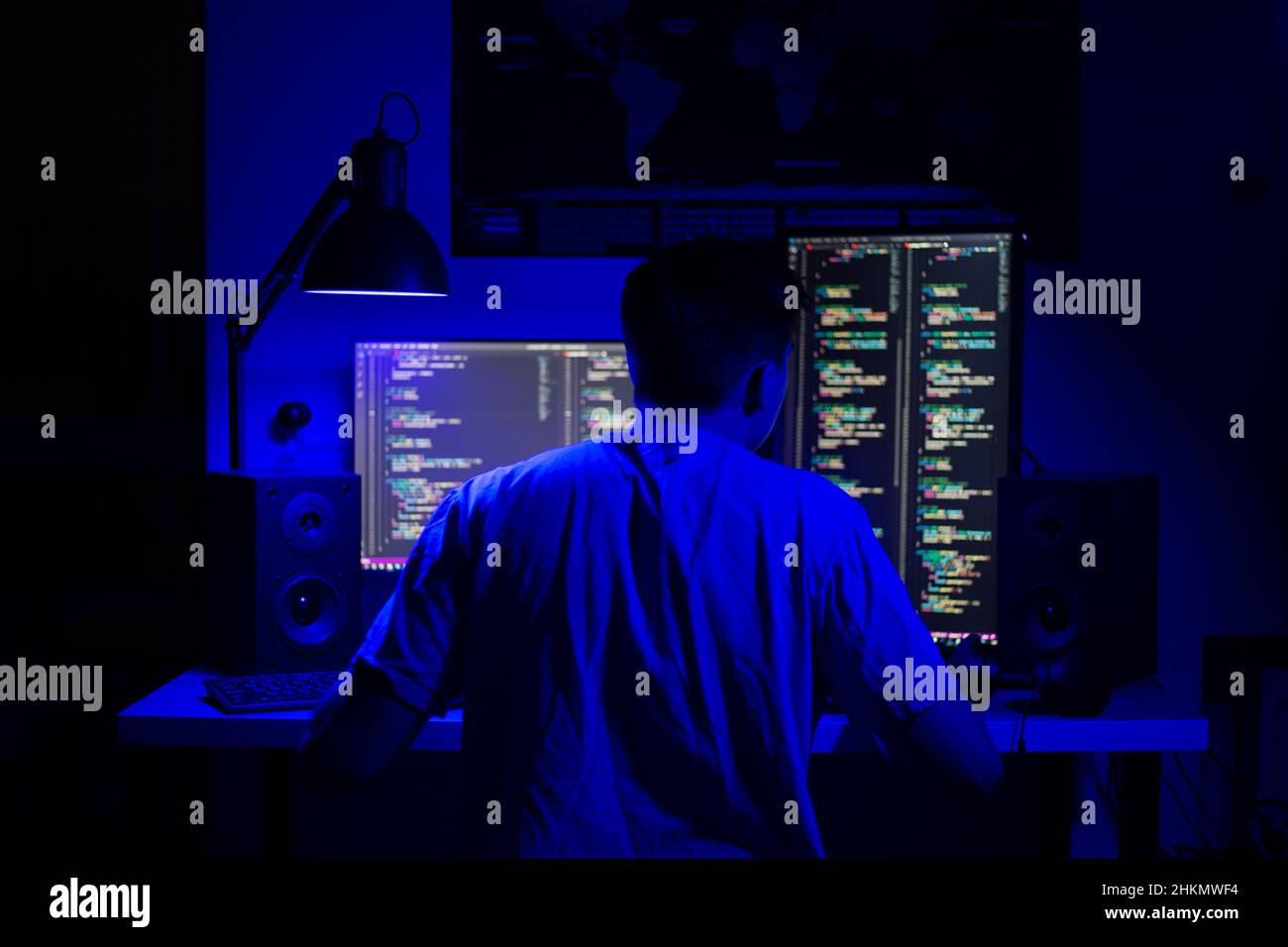 A man sits at a computer in a room at a table at night with blue lighting and programs Stock Photo