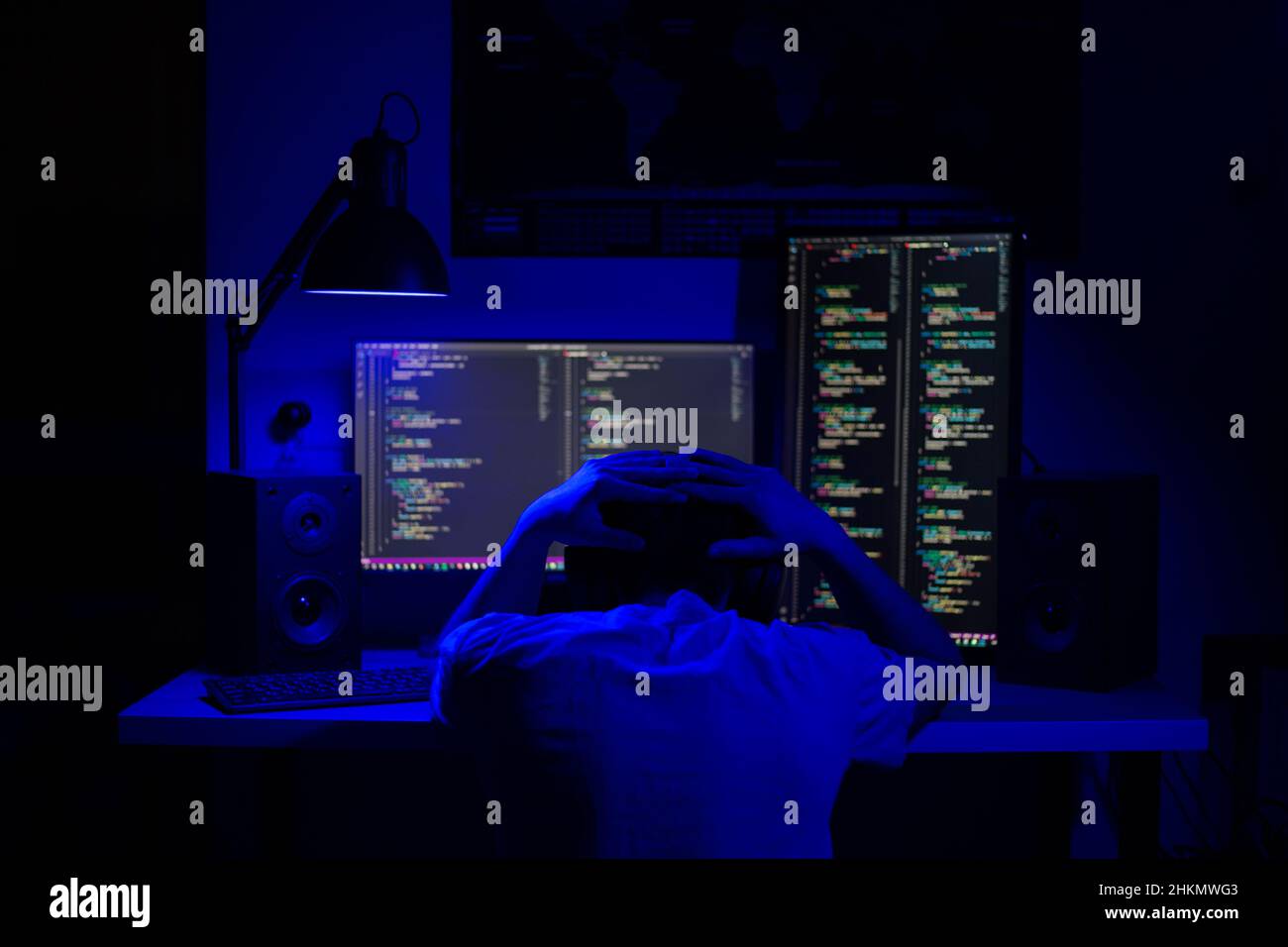 A disappointed man sits at a computer in a room at a table at night with blue lighting and programs Stock Photo