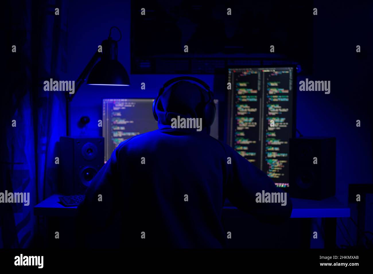 A man in hoodie sits at a computer in a room at a table at night with blue lighting and programs Stock Photo